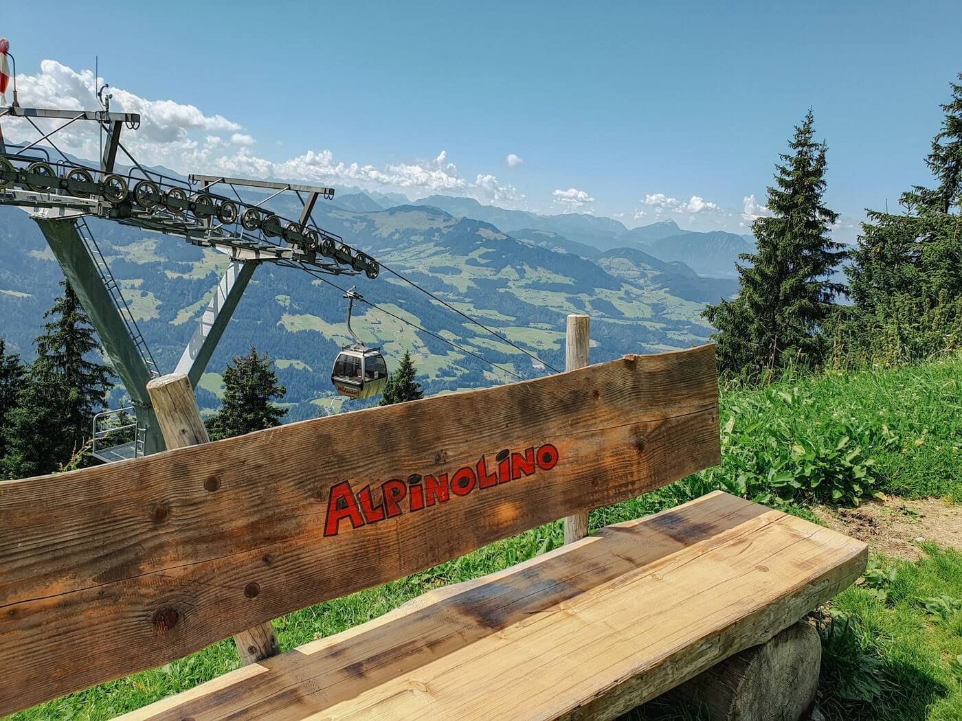 Bank mit Aufschrift Alpinolino vor dem Hintergrund der Bergstation der Alpenrosenbahn in Westendorf