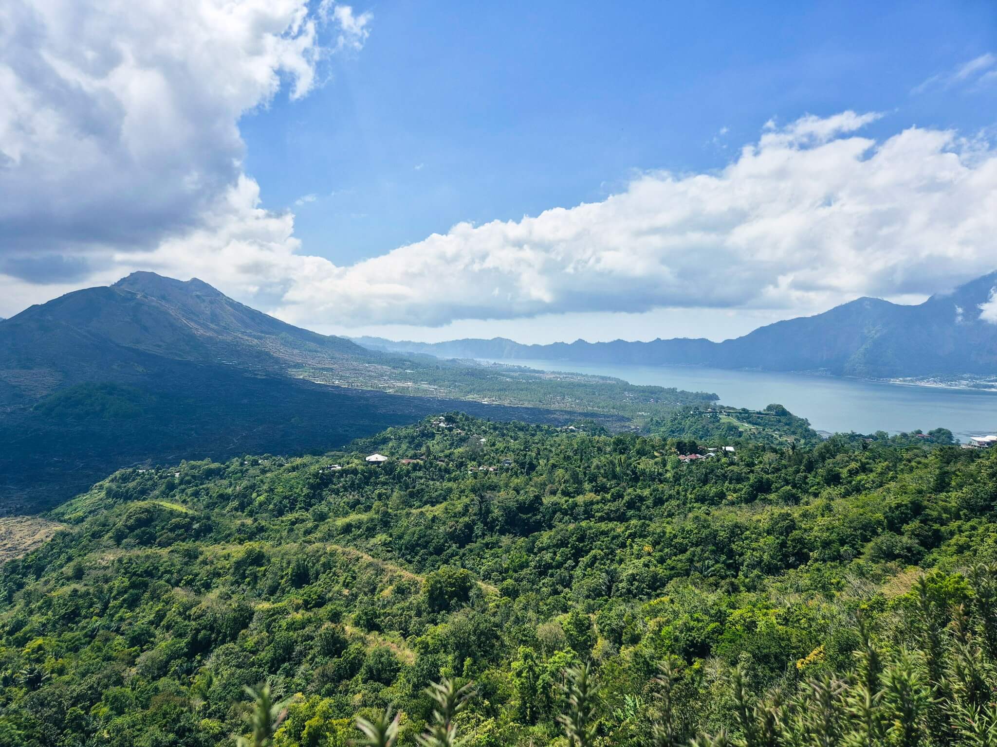 Blick auf den Batursee vor dem Hintergrund des Vulkans Batur und mit viel Grün im Vordergrund